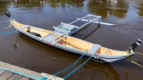 A wooden canoe, built by Micronesian boat builders sits on the water's edge. It is tied to a jetty with blue rope