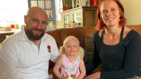 BBC/Seb Cheer A man and woman sit on a sofa, with their three year old daughter between them. They are all smiling at the camera.