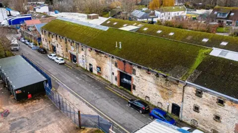 Hollis and Morgan An aerial shot of the building, which is stone and has three levels of windows. The windows are boarded up and the roof has lots of moss growing on it.