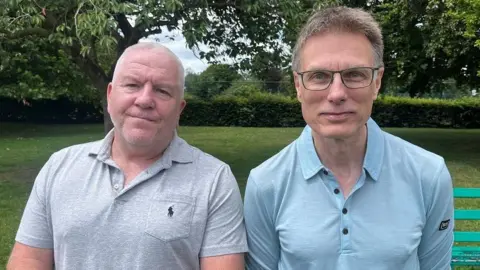 BBC Simon Byrne and Adam Rainford are standing side by side in a park. Simon is wearing a grey polo short while Adam is wearing a blue polo shirt.