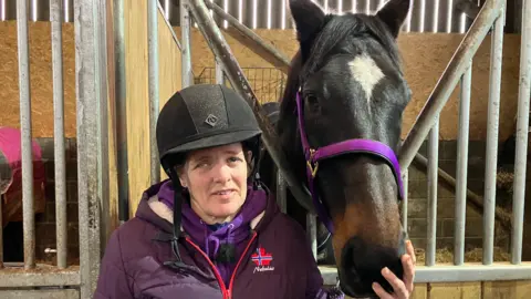 BBC News Lesley Sayers standing in a stable with her horse Seb. She wears a riding cap and purple coat and strokes Seb's nose