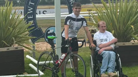 Tom Dunlop is smiling at the camera, wearing cycling gear and holding his bike. While Ollie Meyrick is sitting in his wheelchair and wearing jeans and a white t-shirt which reads "Tom's Big Bike Ride".