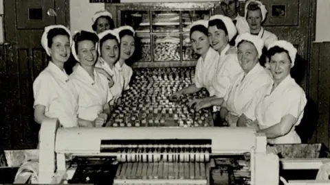 Mars Incorporated Black and white picture of women working at the factory, standing either side of a conveyor belt with chocolate bars on it. They are all wearing white, short-sleeved dresses and white hats with their hair tied up.