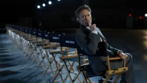 Marvel Robert Downey Junior sitting in a chair in a film studio with his finger over his mouth indicating 'shush'