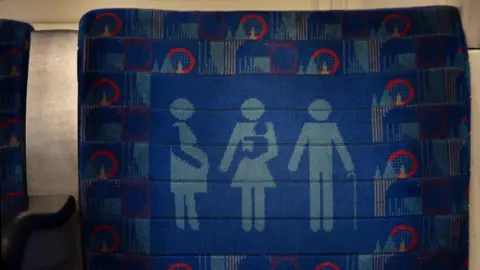 Getty Images Blue fabric upholstery on a Tube seat. The design features icons of a pregnant person, a person holding a baby, and a person with a walking stick, indicating a priority seat.