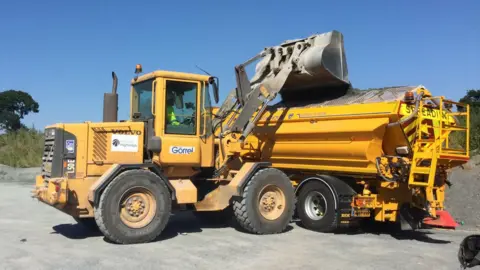 Hampshire County Council A yellow tractor with a scoop bucket refills a yellow gritting lorry at a depot
