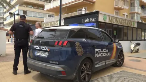 A police car in front of Monaghan's bar. It is blue and there is an officer nearby. He is speaking to a woman.