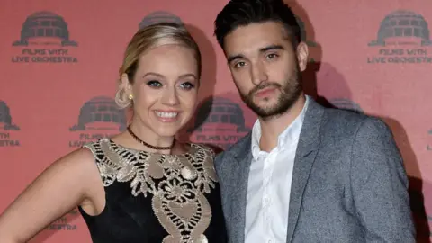 The Wanted singer Tom Parker, wearing a white shirt and grey suit with no tie, with his arm around his smiling blonde wife Kelsey. who is wearing a black and gold dress