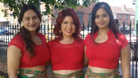 BBC Three women standing, smiling at the camera. They are wearing red tops and green skirts.