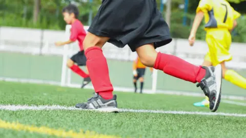 Three children playing sport on an artificial surface. Only the lower half of the children is visible. They are wearing red and yellow sports kit.