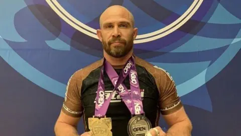 Bea Fernandes/Roots Gym Ben Wyard is wearing a black top with bronze sleeves whilst showing two medals to the camera.