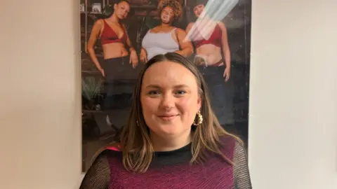 Ellie Brown with long brown hair and gold earrings wearing a black see through top with a hot pink vest underneath. She stands under a poster with three models wearing Recondition clothing. She is smiling.