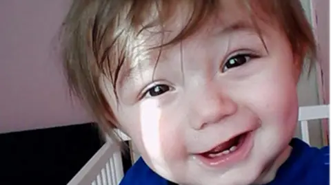 A two-year-old boy in a white cot. He has brown hair sweeping over his forehead, is wearing a blue top and is smiling and looking at the camera.