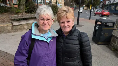 Two women, aged 71 and 65 respectively. Linda on the left, is wearing a purple coat and glasses. Ann, who is on the right, has strawberry blonde hair and a black coat