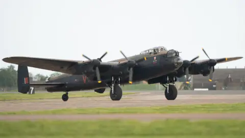 Joe Giddens/PA Wire A large bomber aircraft on the ground with its four propellers spinning. The plane is painted dark green underneath and in camouflage green and brown tones on its upper body. Very short grass is in the foreground of the image.