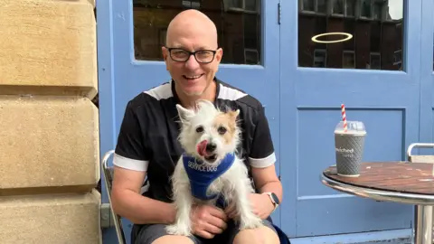 Graham McMillan is wearing black glasses and a black-and-white t-shirt. He is smiling at the camera and holding Maisie, a white and beige Jack Russell and Yorkie cross. She is licking her nose and wearing a blue harness that reads 'Service Dog'. Behind them is part of a sandstone wall and blue double-doors on the side of Bewiched Coffee in Peterborough. 