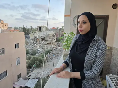 BBC Hanya, who is wearing a black headscarf, looks out at a street filled with destroyed buildings, from her balcony