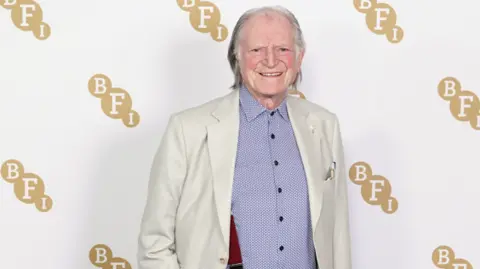 PA Media David Bradley pictured smiling against a white background with the letters BFI in gold. Bradley is wearing a blue shirt under a white suit jacket and red braces.