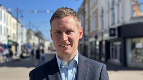 James is standing in the centre of Coleraine with shops behind him. He has fair hair and is wearing a navy blazer and blue shirt. He is looking directly at the camera and smiling