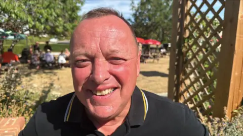 Richard Knights/BBC A man is smiling, sat outside in a busy and sunny pub garden