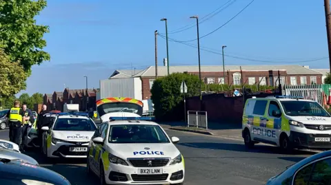 A number of police cars parked at the side of a road. Officers can be seen talking to each other on a pavement on the left.
