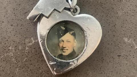 A heart-shaped silver pendant on a pin with a circular frame containing an old black and white photograph of a man smiling and wearing a garrison cap with a military badge on the side