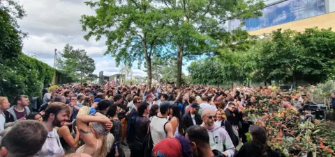 Joseph Winters Dozens of people stand under a tree beside a building on a summer's day