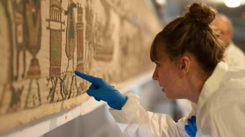 French Ministry of Culture: A woman in a white hazmat suit and blue gloves places a finger on the tapestry.