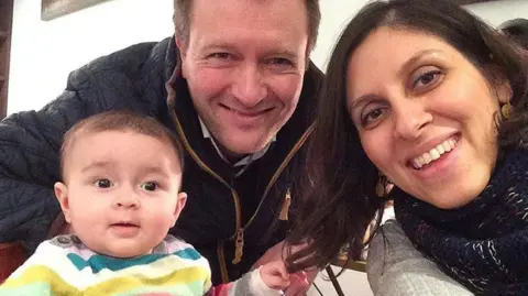 Family photo A selfie image of a man, a woman and a baby. The man is wearing a navy jacket and brown hair. A woman has dark hair and is wearing a grey jumper. The baby is wearing a multicoloured striped top and a little bit of dark hair.