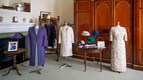 Royal Collection Trust Four headless mannequins wearing some of the late Queen's clothes, exhibited in a room with wooden furniture, a fireplace, a hat rack holding three hats on a table which is also holding hand bags and an umbrella.