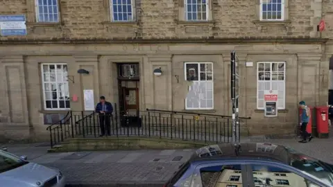 Brown brick building which says Post Office. Man stands outside. Two cars parked on the road outside. 