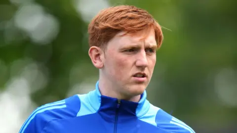 Bradley Collyer/PA Wire Angry Ginge. Young man with ginger hair wears a blue Soccer Aid top. The photos has a blurry green background. 