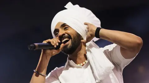 Getty Images Singer Diljit Dosanjh wearing all white traditional Punjabi attire including a white Turban (religious headwear), while singing into a black microphone, with his left hand touching his head. The background is dark with a spotlight.