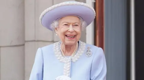 Getty Images The late Queen Elizabeth II smiles broadly as she stands outside, wearing a pale blue hat and jacket; her white gloves can just be seen and she has several strands of pearls around her neck