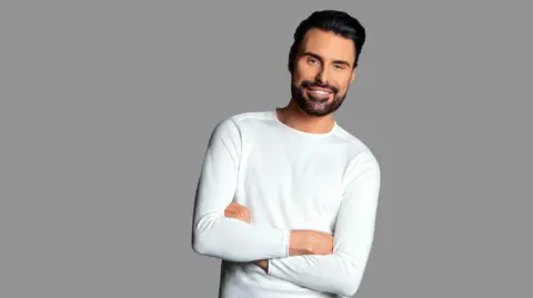 Rylan Clark-Neal stands with his arms crossed - he has short black hair and a beard