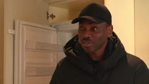 Chinedu Nwoye is wearing a black cap and dark jacket, and is standing in front of an open fridge.