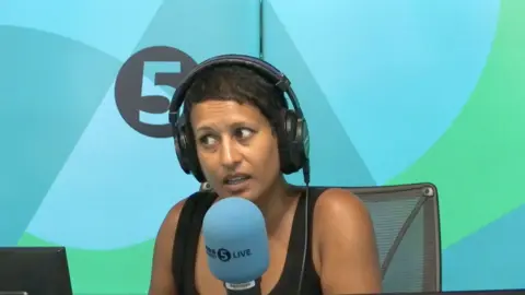 Naga Munchetty has short black hair. She is wearing a black vest top and is sitting behind a 5 Live microphone.