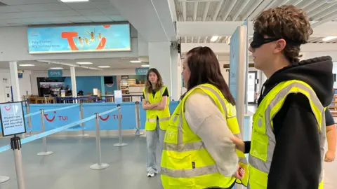 A couple in yellow safety jackets role play guiding a visually impaired person around Exeter airport. In the background you can see a Tui check in desk and a trainer from Guide Dogs. The person being guided wears a black blindfold.