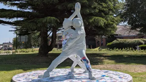 The grey sculpture, which is two people intertwined with their hands joined above their heads. Colourful patterns can be seen in sections through the sculpture. It is on a patch of grass with a road behind it.