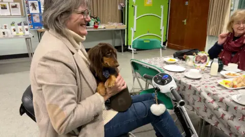 An older woman smiles as she holds her brown and tan dachshund on her lap and sits on her mobility scooter at a table filled with party food in a community centre. Another older woman in a red scarf is sat opposite her.