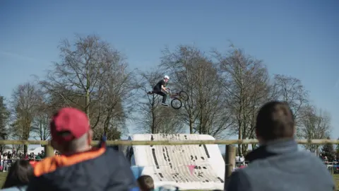 A BMX rider performing a stunt after jumping from a white ramp. The rider, wearing black clothes and a white helmet, appears suspended in the air, holding his bike in front of him. The backs of people's heads can be seen as they watch. Tall bare trees can be seen under a blue sky.
