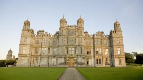 BBC The front of Burghley House, with a pathway leading up to a golden entrance and a neat lawn either side. The building is very grandeur, with several windows on three floors as well as turrets.