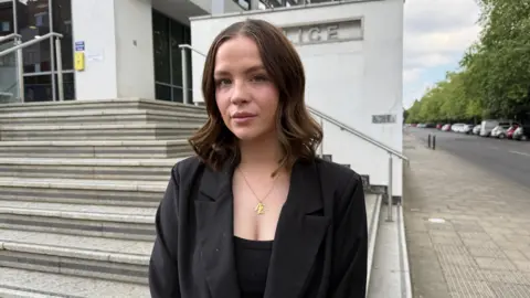 A young woman with black blazer and black top. She is standing in front of Warwick Justice Centre and has shoulder length brown hair.