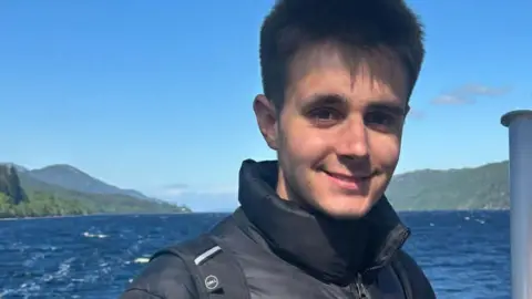 A head and shoulders image of James Thompson. A young man wearing a black puffer jacket with two black rucksack straps over it. He appears to be on a boat, which has Union Jack flag on it. There is water and mountains in the background.