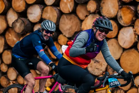 Red On Sports A woman on a bike gives a thumbs up, she is in a multi-coloured top with a black and pink helmet. Behind her is a man with a beard on a pink bike with a silver helmet. They are cycling past a lot of chopped wood.