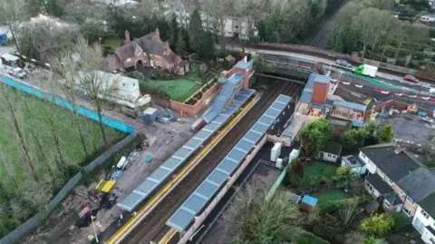 WMCA An aerial view of Pineapple Road station in Kings Heath, shows the station platforms on either side of the railway linem as well as a road bridge going over the line. Green spaces with properties on them can be seen on either side of the station. 