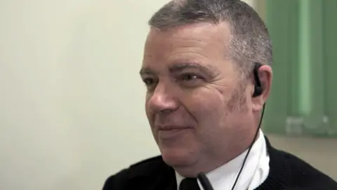 Head and shoulders image of John Sneddon who is looking off to one side. He has cropped, grey hair and is wearing a white shirt, black tie and black jumper. He also has an earpiece in.