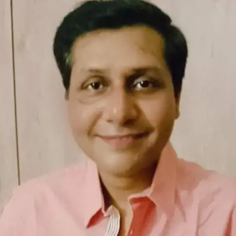 Sapna Mukherjee Sougat smiling at the camera. He has dark hair and is wearing a pink shirt.