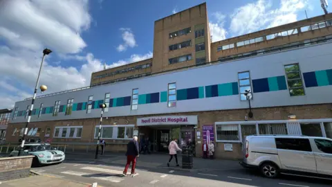 People walking into Yeovil District Hospital. The building has brown bricks and purple lettering saying 'Yeovil District Hospital'. There are cars outside and a zebra crossing.