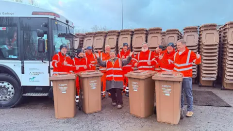 Herefordshire Council A group of refuge workers in orange high viz jackets stand around brown plastic waste bins with a white refuge vehicle on the left. 